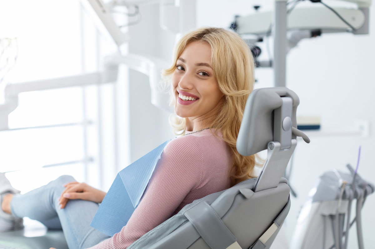 Smiling and relaxed young woman sitting at dental chair in modern clinic, waiting for her dentist. Happy attractive blonde lady attending luxury dental clinic, bacl view, copy space